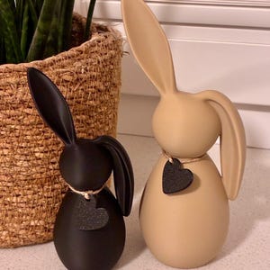 May include: Two decorative bunny figurines. One is black, the other is tan. Each has long ears and a heart-shaped tag. The black bunny has a white string around its neck. The tan bunny has a brown string around its neck. A woven basket with a plant is in the background.
