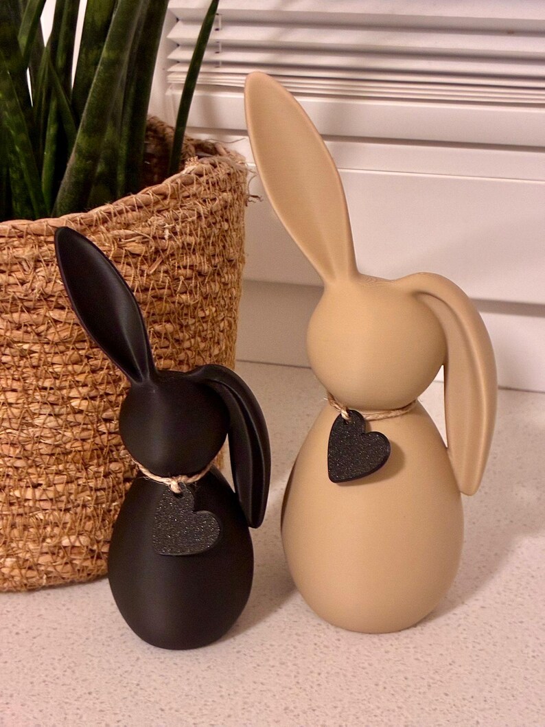 May include: Two decorative bunny figurines. One is black, the other is tan. Each has long ears and a heart-shaped tag. The black bunny has a white string around its neck. The tan bunny has a brown string around its neck. A woven basket with a plant is in the background.