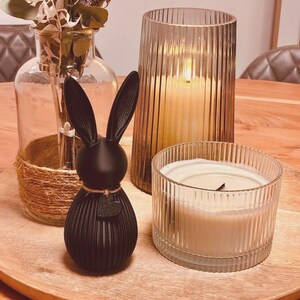 May include: A decorative wooden tray holds a black bunny figurine, two glass candle holders, and a vase with dried flowers. The bunny has a textured, ribbed design. The candles are lit, casting a warm glow. The vase is wrapped with twine.