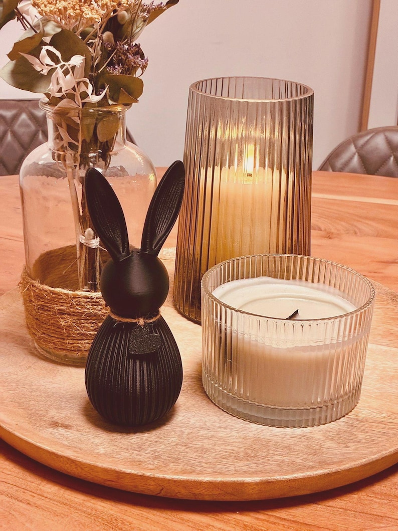 May include: A decorative wooden tray holds a black bunny figurine, two glass candle holders, and a vase with dried flowers. The bunny has a textured, ribbed design. The candles are lit, casting a warm glow. The vase is wrapped with twine.