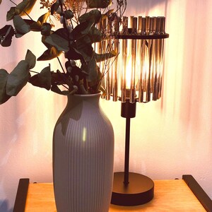 May include: A decorative table lamp with a cylindrical crystal shade and a black base. A gray vase with vertical ridges holds dried flowers and greenery. The lamp and vase sit on a wooden surface, creating a warm, inviting ambiance.