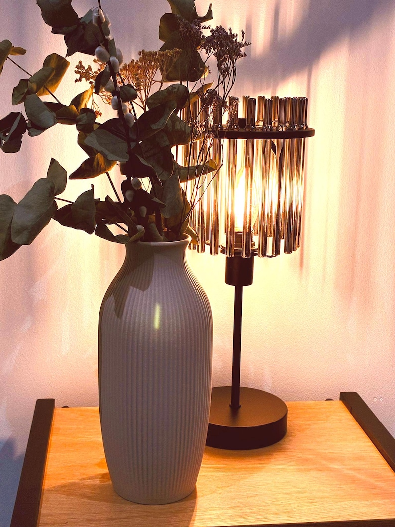 May include: A decorative table lamp with a cylindrical crystal shade and a black base. A gray vase with vertical ridges holds dried flowers and greenery. The lamp and vase sit on a wooden surface, creating a warm, inviting ambiance.