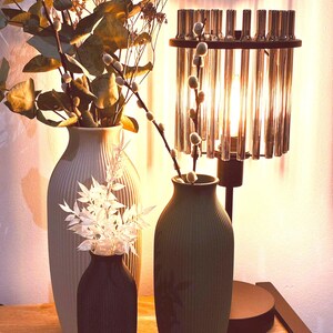 May include: A decorative arrangement featuring three vases of varying sizes and colors, holding dried flowers and branches. A modern lamp with a cylindrical glass shade is also present, creating a warm, inviting ambiance. The vases are in shades of gray, black, and green.