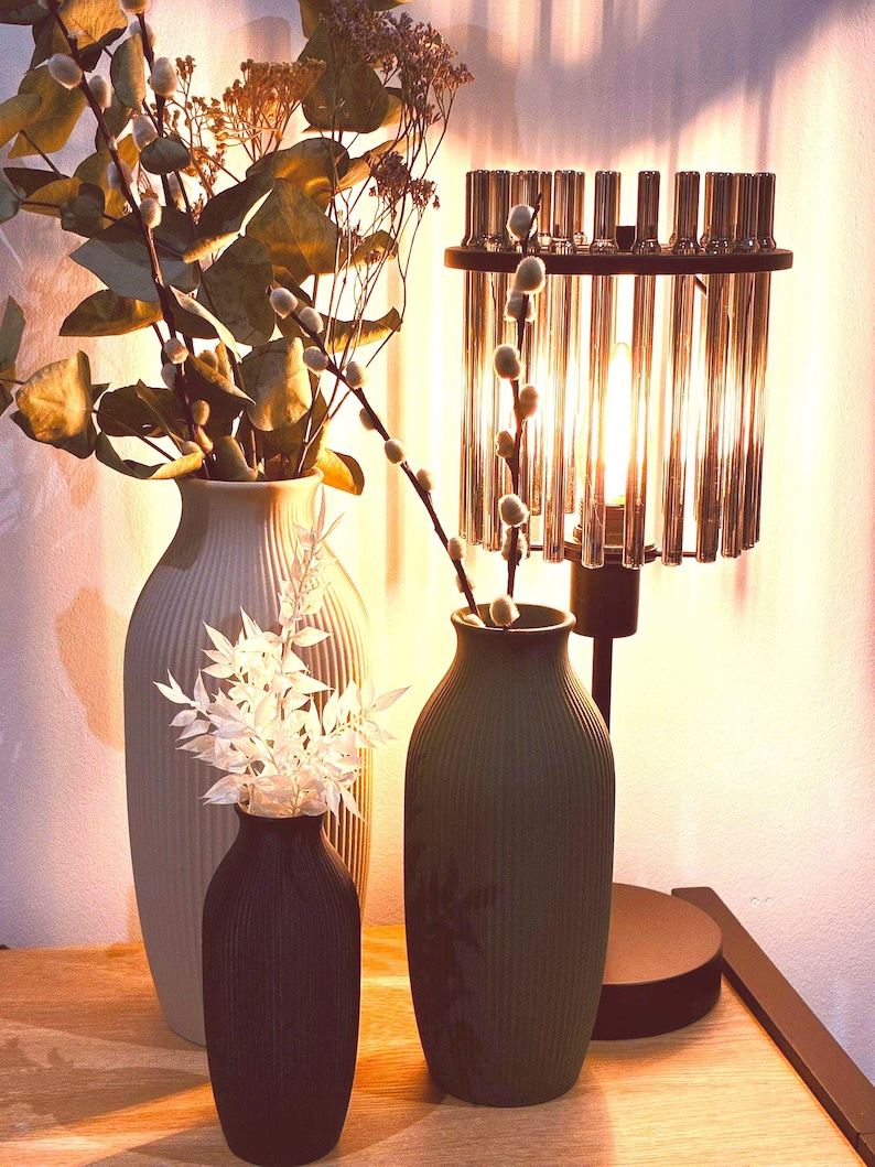 May include: A decorative arrangement featuring three vases of varying sizes and colors, holding dried flowers and branches. A modern lamp with a cylindrical glass shade is also present, creating a warm, inviting ambiance. The vases are in shades of gray, black, and green.