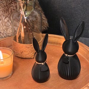 May include: Two black rabbit figurines with long ears and textured bodies sit on a wooden tray. Each rabbit has a small heart-shaped detail and is adorned with twine. A lit candle and a glass vase with dried flowers are also on the tray.