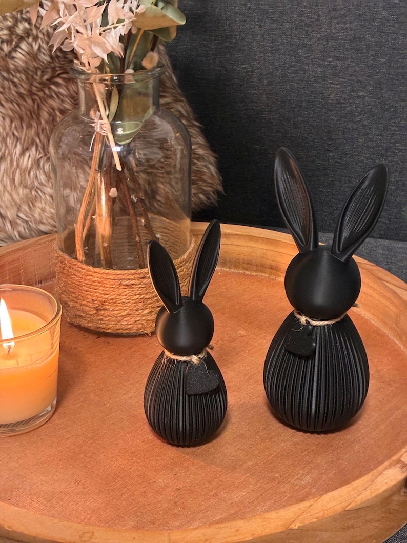 May include: Two black rabbit figurines with long ears and textured bodies sit on a wooden tray. Each rabbit has a small heart-shaped detail and is adorned with twine. A lit candle and a glass vase with dried flowers are also on the tray.