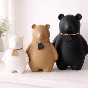 Pu&ograve; includere: Tre statuette di orsi in ceramica, bianche, beige e nere. Ogni orso ha un'etichetta a forma di cuore intorno al collo. Gli orsi sono esposti su una mensola bianca, mostrando un design minimalista.