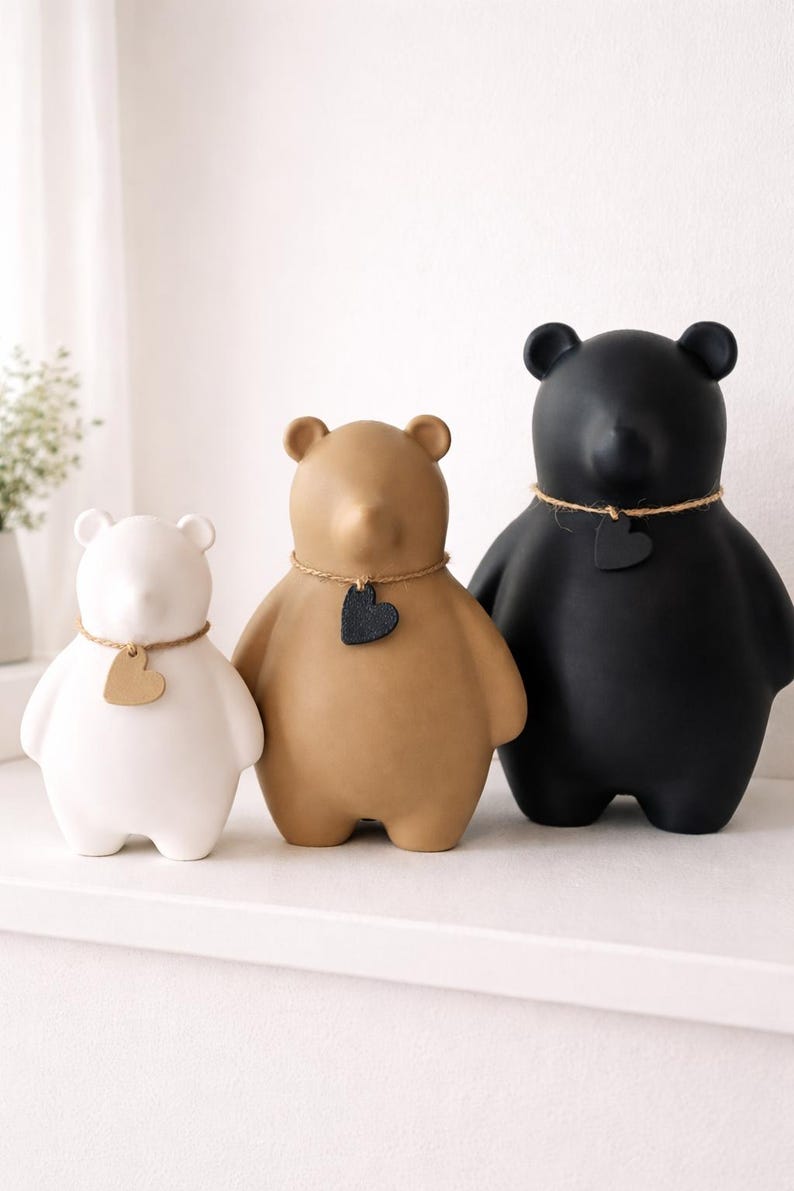 Pu&ograve; includere: Tre statuette di orsi in ceramica, bianche, beige e nere. Ogni orso ha un'etichetta a forma di cuore intorno al collo. Gli orsi sono esposti su una mensola bianca, mostrando un design minimalista.