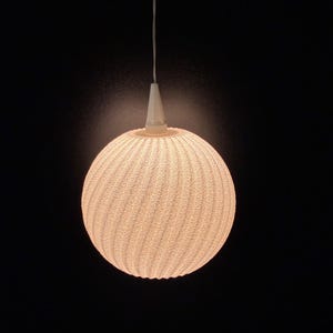 Puede incluir: Una l&aacute;mpara colgante esf&eacute;rica blanca con una superficie texturizada y acanalada. La luz cuelga de un cable blanco y un accesorio en forma de cono sobre un fondo oscuro. La luz est&aacute; encendida, creando un brillo c&aacute;lido.