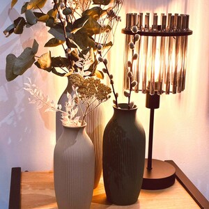 May include: A decorative arrangement featuring three ceramic vases in neutral tones, with vertical ribbed detailing. The vases hold dried flowers and branches. A crystal lamp with a black base is to the right, casting a warm glow.