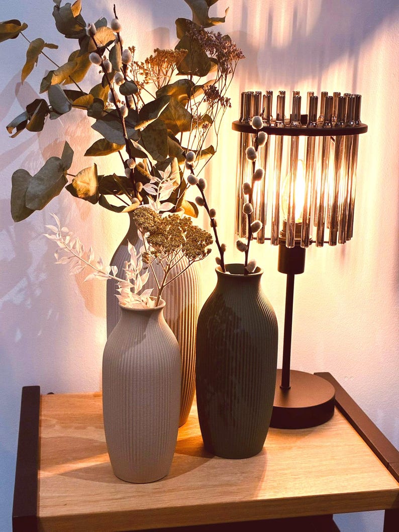 May include: A decorative arrangement featuring three ceramic vases in neutral tones, with vertical ribbed detailing. The vases hold dried flowers and branches. A crystal lamp with a black base is to the right, casting a warm glow.