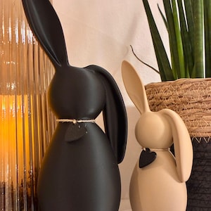 May include: Two decorative bunny figurines. One is matte black, the other is beige. Each has long ears, a rounded body, and a small black heart pendant. They are tied with twine around their necks.