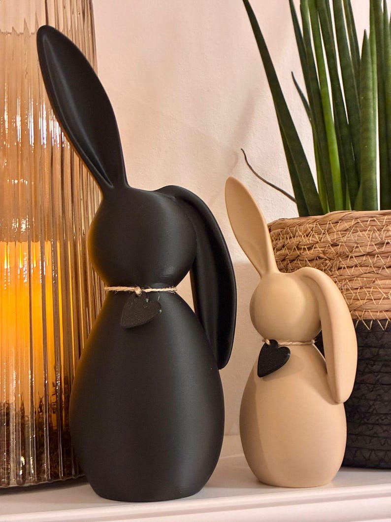 May include: Two decorative bunny figurines. One is matte black, the other is beige. Each has long ears, a rounded body, and a small black heart pendant. They are tied with twine around their necks.