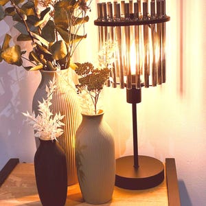 May include: A decorative table lamp with a black base and a cylindrical shade composed of vertical glass rods. Three vases of varying sizes and colors hold dried flowers and foliage. The scene is set on a wooden surface.