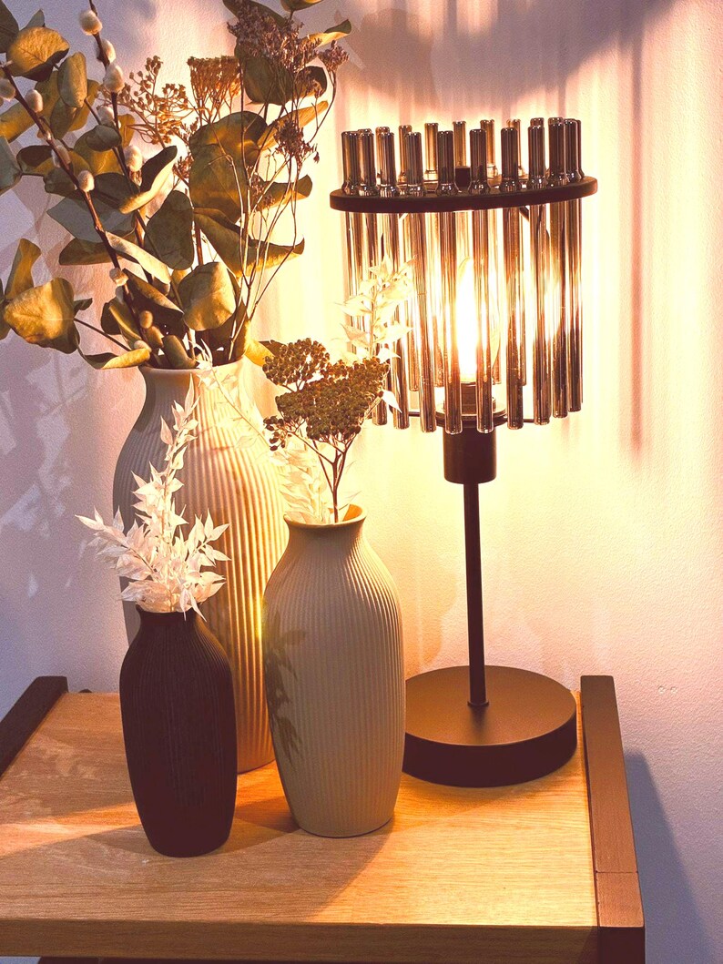 May include: A decorative table lamp with a black base and a cylindrical shade composed of vertical glass rods. Three vases of varying sizes and colors hold dried flowers and foliage. The scene is set on a wooden surface.