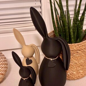 May include: Three decorative bunny figurines in varying sizes and colors. The largest is black, the middle one is tan, and the smallest is black. Each bunny has long ears and a heart-shaped accent. The bunnies are placed on a white surface.