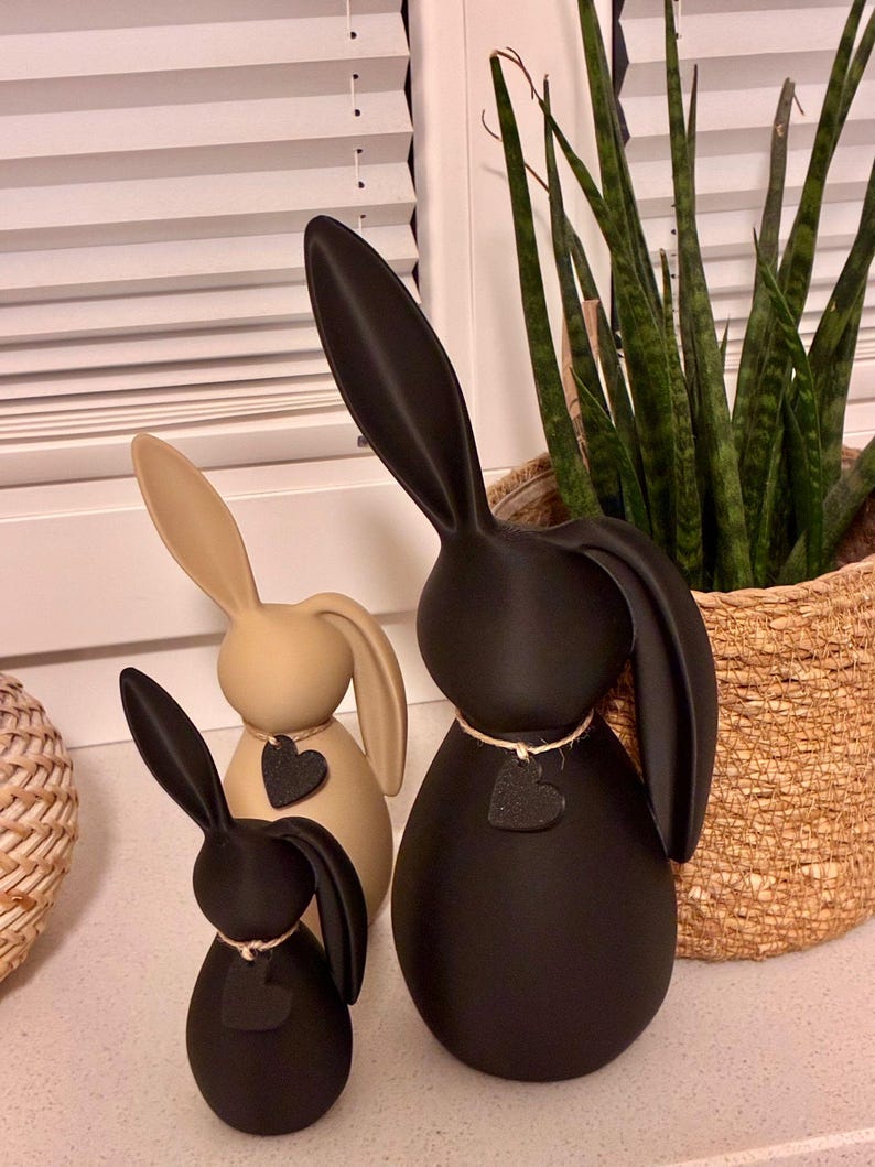 May include: Three decorative bunny figurines in varying sizes and colors. The largest is black, the middle one is tan, and the smallest is black. Each bunny has long ears and a heart-shaped accent. The bunnies are placed on a white surface.