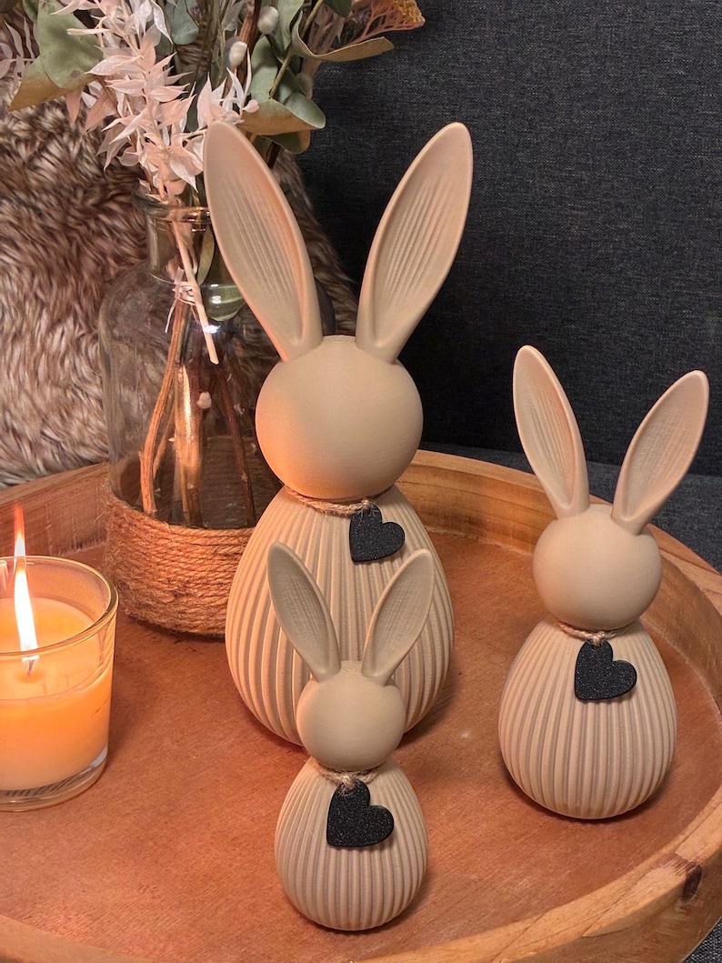 May include: Three beige bunny figurines of varying sizes, each with long ears and a black heart pendant. The bunnies are on a wooden tray, with a lit candle and a vase of flowers in the background. The figurines have a ribbed texture.
