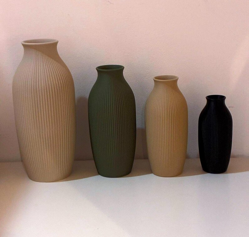 May include: Four ceramic vases in varying sizes and colors. The largest is beige, the next is olive green, followed by a smaller beige vase, and the smallest is black. All vases have vertical ribbed detailing.
