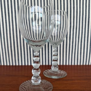 May include: Two clear wine glasses with a bubble-style stem. The glasses are on a wooden surface with a striped blue and white background. The glasses are empty and reflect the background.