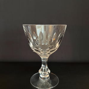 May include: A clear, cut-glass coupe glass with a wide bowl and a short stem. The glass features a repeating pattern of oval shapes around the bowl. The base is a flat, circular disc. The glass is designed for serving cocktails or champagne.
