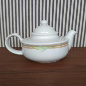 Puede incluir: Tetera de cerámica blanca con tapa y asa. La tetera tiene una banda decorativa amarilla y verde cerca de la base. El fondo es un patrón de rayas blancas y grises.