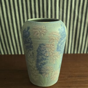 May include: A cylindrical ceramic vase with a speckled light blue base color. The exterior surface features a repeating floral pattern in shades of soft pink and deep blue, depicting clusters of blossoms that wrap around the body of the vessel.