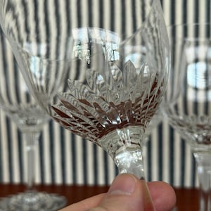 Puede incluir: Copas de vino de cristal transparente con un detallado diseño floral en la copa. El tallo es delgado y elegante. Las copas se sostienen en una mano, con un fondo a rayas.