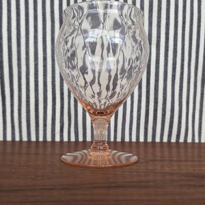Puede incluir: Una copa de cristal teñida de rosa con un patrón texturizado en forma de diamante en el cuenco. La copa tiene un tallo delgado y una base circular. El fondo presenta una tela a rayas, y la copa se encuentra sobre una superficie de madera oscura. Cristalería elegante.