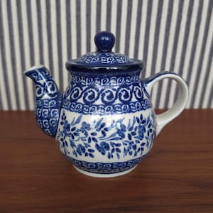 Puede incluir: Una pequeña tetera de cerámica azul y blanca con tapa. La tetera presenta un estampado floral y un pico curvo. El asa es blanca. La tetera mide aproximadamente 13 cm de altura.
