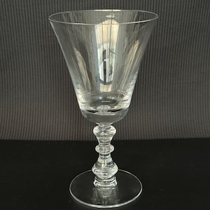 Puede incluir: Una copa de vino transparente con un cuenco ancho y cónico y un tallo decorativo en forma de balaustre. La copa se asienta sobre una base circular. La copa es de cristal transparente y refleja la luz. Altura aproximada 18 cm.