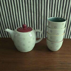 May include: A ceramic teapot with a maroon lid and a light-colored body featuring small gray circular patterns, accompanied by a stack of four matching ceramic cups, each with a light-colored exterior and a distinct interior color rim.