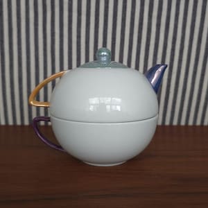 May include: A white ceramic teapot and teacup set. The teapot has a blue spout and a green lid with a decorative knob. The teacup has a purple handle, and the teapot has a gold handle. The set is on a dark wooden surface.