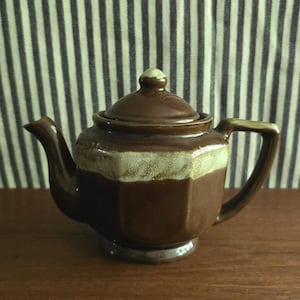 May include: A small, hexagonal ceramic teapot with a glossy dark brown glaze and a contrasting cream-colored band around the upper body. The teapot features a matching lid with a small knob, a curved spout, and a sturdy handle on the side.