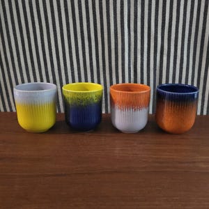 Puede incluir: Cuatro tazas de cerámica con textura acanalada vertical y acabado de esmalte de goteo en dos tonos. La primera taza es gris claro y amarilla, la segunda es amarilla y azul oscuro, la tercera es naranja y gris claro, y la cuarta es azul oscuro y naranja.
