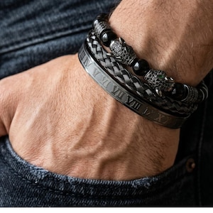 Könnte beinhalten: Eine Sammlung schwarzer Armbänder an einem Handgelenk. Die Armbänder umfassen ein geflochtenes Design, ein Band mit römischen Ziffern und ein Perlenarmband mit Pantherkopf-Akzenten. Die Pantherköpfe haben grüne Augen.