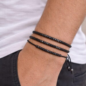 Può includere: Tre braccialetti neri con perline sono indossati su un polso. I braccialetti sono fatti di piccole perline quadrate infilate su cordoncini neri. I braccialetti sono regolabili e hanno un design minimalista. Il polso è contro una camicia bianca.
