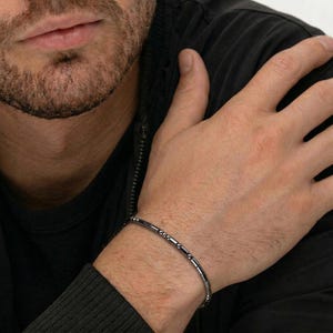 May include: A dark grey beaded bracelet featuring cylindrical and rounded beads. The bracelet is worn on a wrist, against a black jacket background.