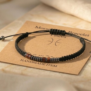 Puede incluir: Pulsera de macramé negra con cuentas de madera y metálicas. La pulsera está sobre una tarjeta que dice "Pulsera Morse Piscis" y "Artículo hecho a mano". La pulsera es ajustable.