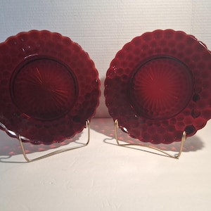 May include: Two matching round plates made of deep red translucent glass. Each plate features a raised hobnail texture along the outer rim and a scalloped edge. The center of each plate displays a sunburst pattern radiating from the middle.