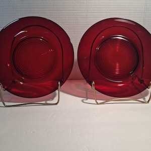 May include: Two circular ruby red glass plates featuring a concentric ring pattern in the center. Each plate is displayed upright on a simple gold-toned wire stand, showcasing the translucent quality and deep red color of the pressed glass material.