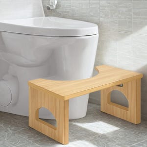 May include: A light-colored wooden toilet stool with a curved top and cut-out sides. The stool is positioned in front of a white toilet, providing a helpful aid for users. The stool is made of wood and has a natural finish.