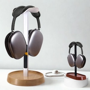 Puede incluir: Auriculares grises sobre la oreja exhibidos en un soporte de madera y metal. El soporte principal tiene una base de madera circular y un soporte vertical de metal. También se ve un soporte más pequeño.
