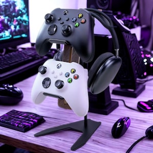 Op de afbeelding: Een zwart-witte gaming controller stand die twee controllers en een headset vasthoudt. De standaard is zwart met een houten steun. De controllers zijn zwart en wit met kleurrijke knoppen. De headset is zwart.