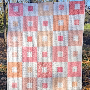 May include: A handmade quilt with a patchwork design. The quilt features a grid pattern of squares in shades of pink, peach, and white. The squares have various textures and patterns, creating a visually appealing and cozy aesthetic. The quilt is hanging outdoors.