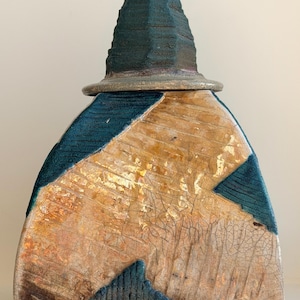 Può includere: Un vaso in ceramica fatto a mano con una combinazione di colori verde acqua, beige e oro. Il vaso ha un corpo triangolare strutturato con un coperchio conico. Il design include forme geometriche verde acqua e una finitura craquelé. Il vaso è sostenuto da tre piccoli piedi.