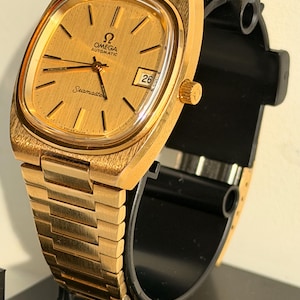 Könnte beinhalten: Eine goldfarbene Omega Seamaster Automatik-Armbanduhr. Die Uhr hat ein rechteckiges Zifferblatt mit Datumsanzeige. Das Armband und das Zifferblatt sind goldfarben. Die Uhr wird auf einem schwarzen Ständer präsentiert.