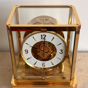 May include: A gold-toned Jaeger-LeCoultre Atmos clock encased in a rectangular glass cabinet. The white circular dial features gold-coloured Arabic numerals at 3, 6, 9, and 12, with gold hands and visible internal mechanical gears behind the face.