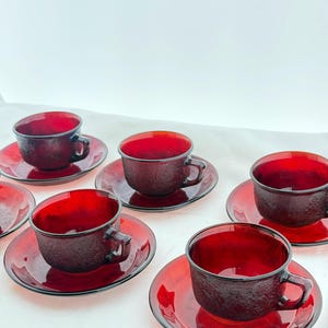 Può includere: Set di sei tazze da tè e piattini in vetro rosso rubino. Ogni tazza ha un esterno strutturato e un manico. I piattini sono rotondi con un bordo leggermente rialzato. Il set è disposto su una superficie bianca.