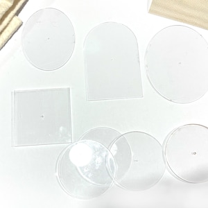 May include: Clear acrylic shapes in various forms, including circles, a square, and an arched shape. The shapes are arranged on a white surface, with a beige fabric in the background.